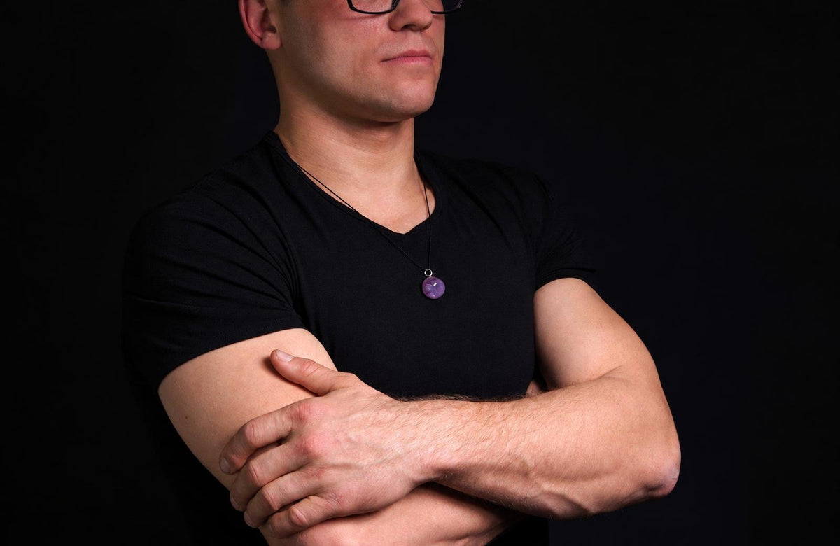 Vyras juodu marškinėliu dėvi kaklo apyrankę su violetiniu, apvaliu pakabuku ant tamsaus fono.