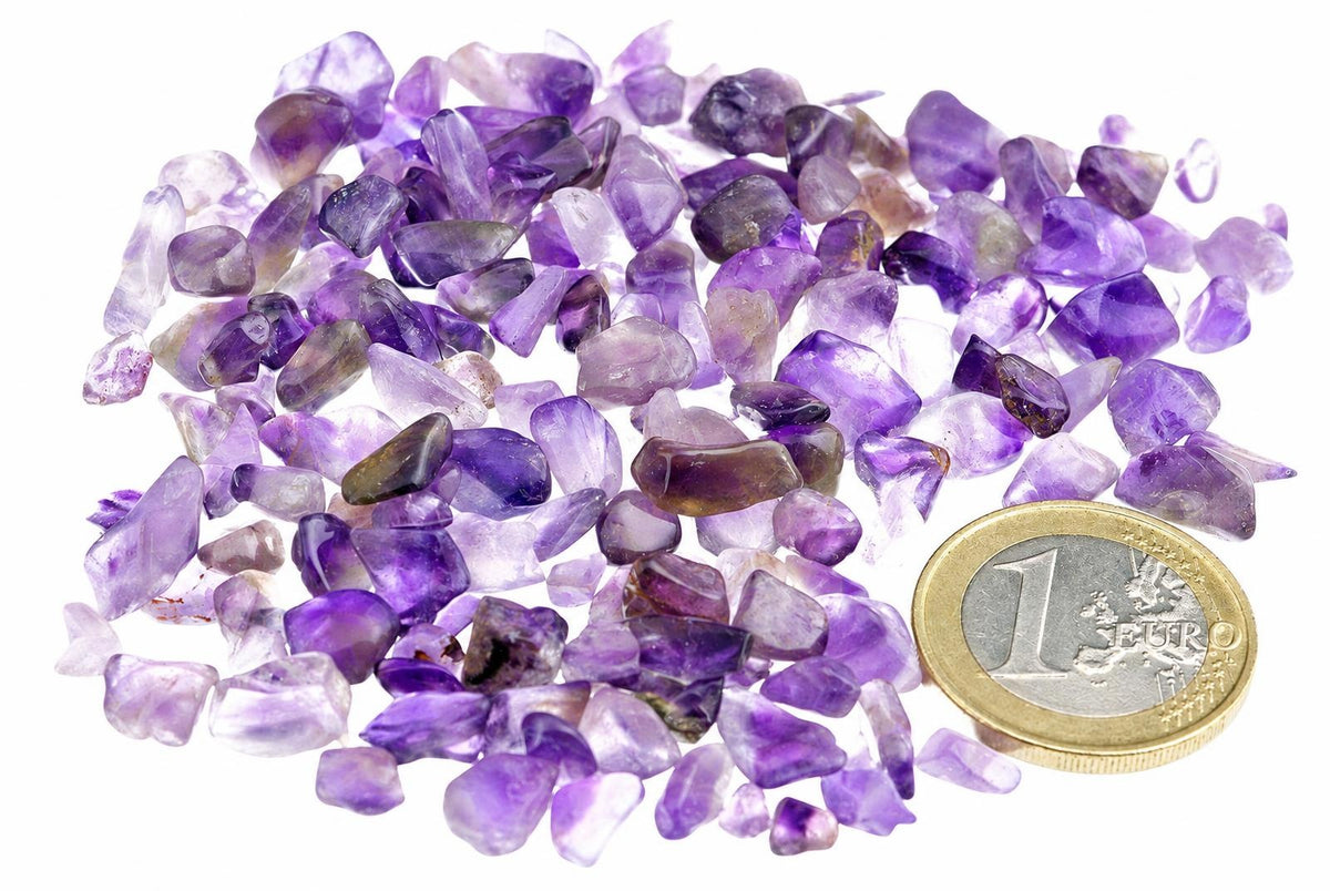 Pile of small, rough amethyst stones in shades of purple and violet on a white surface next to a one euro coin.