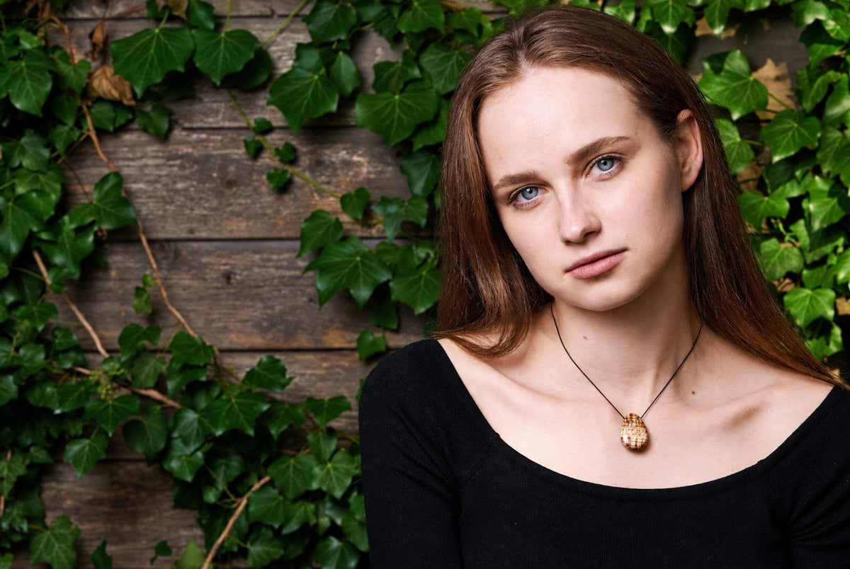 Moteris su ilgomis raudonais plaukais ir mėlynomis akimis, dėvinti juodą marškinėlę ir auksinį pakabuką ant kaklo, prieš tamsų medinį foną su žaliais vijokliniais augalais.