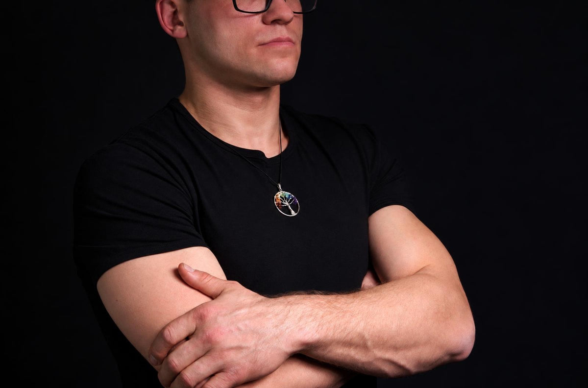Vyras juodu marškinėliu dėvi pakabuką su spalvotu medžio siluetu apskritame rėme ant tamsaus fono.
