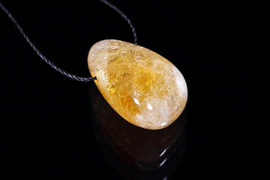 Geltonas, poliruotas citrino akmuo ant juodos virvelės pakabukas tamsiame fone.