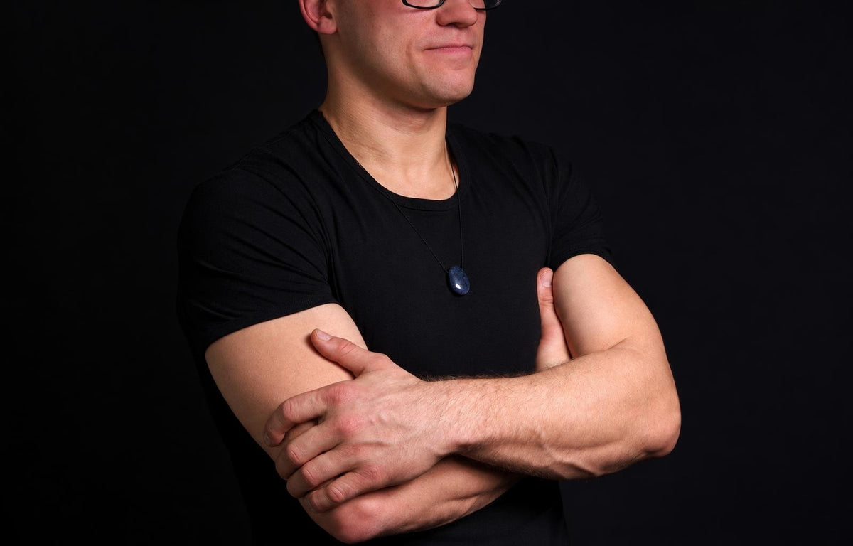 Vyras juodu marškinėliu su tamsiai mėlynu ovaliu pakabuku kaklo grandinėje, rankos susikryžusios, tamsi fona.