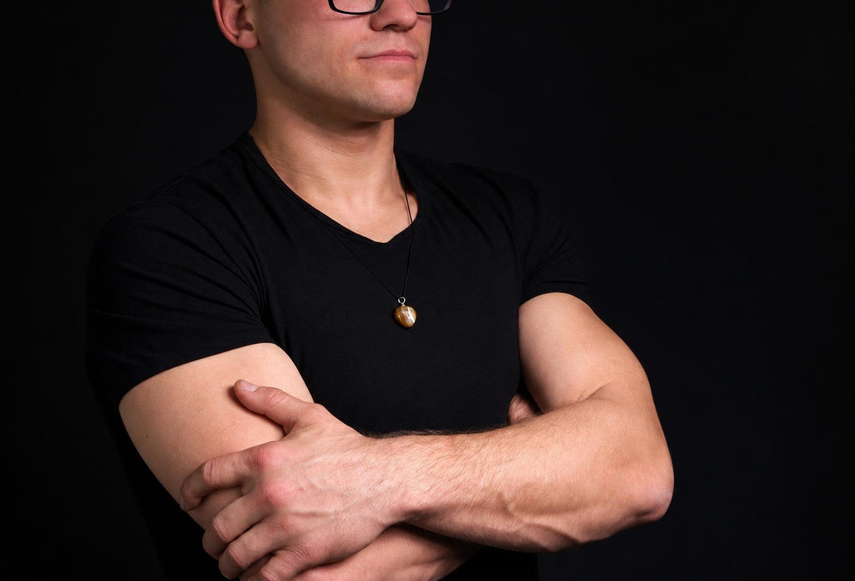 Vyras juodu marškinėliu su tamsiai ruda, ovališka pakabuka ant kaklo prieš tamsų foną.