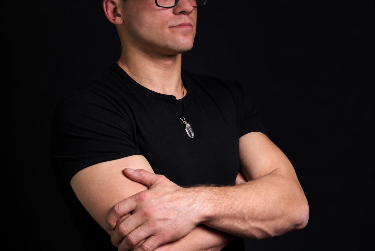 Vyras juodu marškinėliu dėvi pakabuką su skaidriu, akmens formos akmenuku ant tamsaus fono.