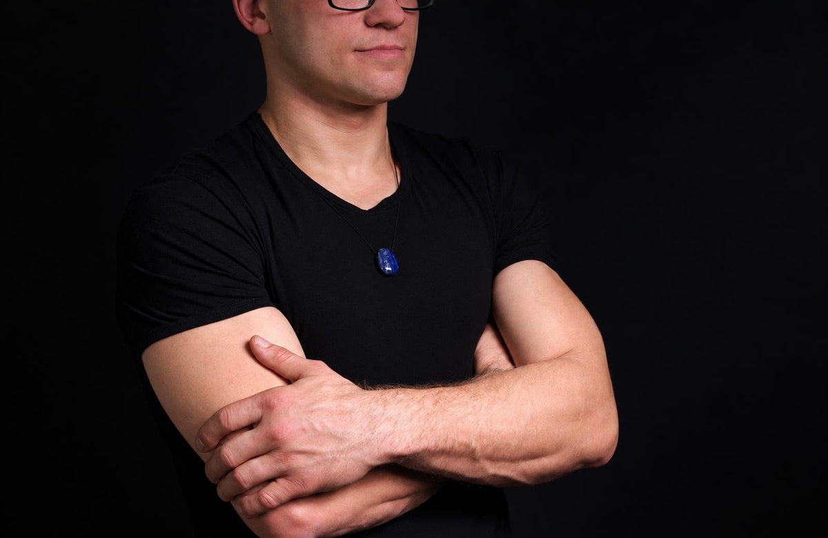 Vyras juodu marškinėliu dėvi pakabuką su tamsiai mėlynu, ovaliu lazurito akmeniu ant kaklo.