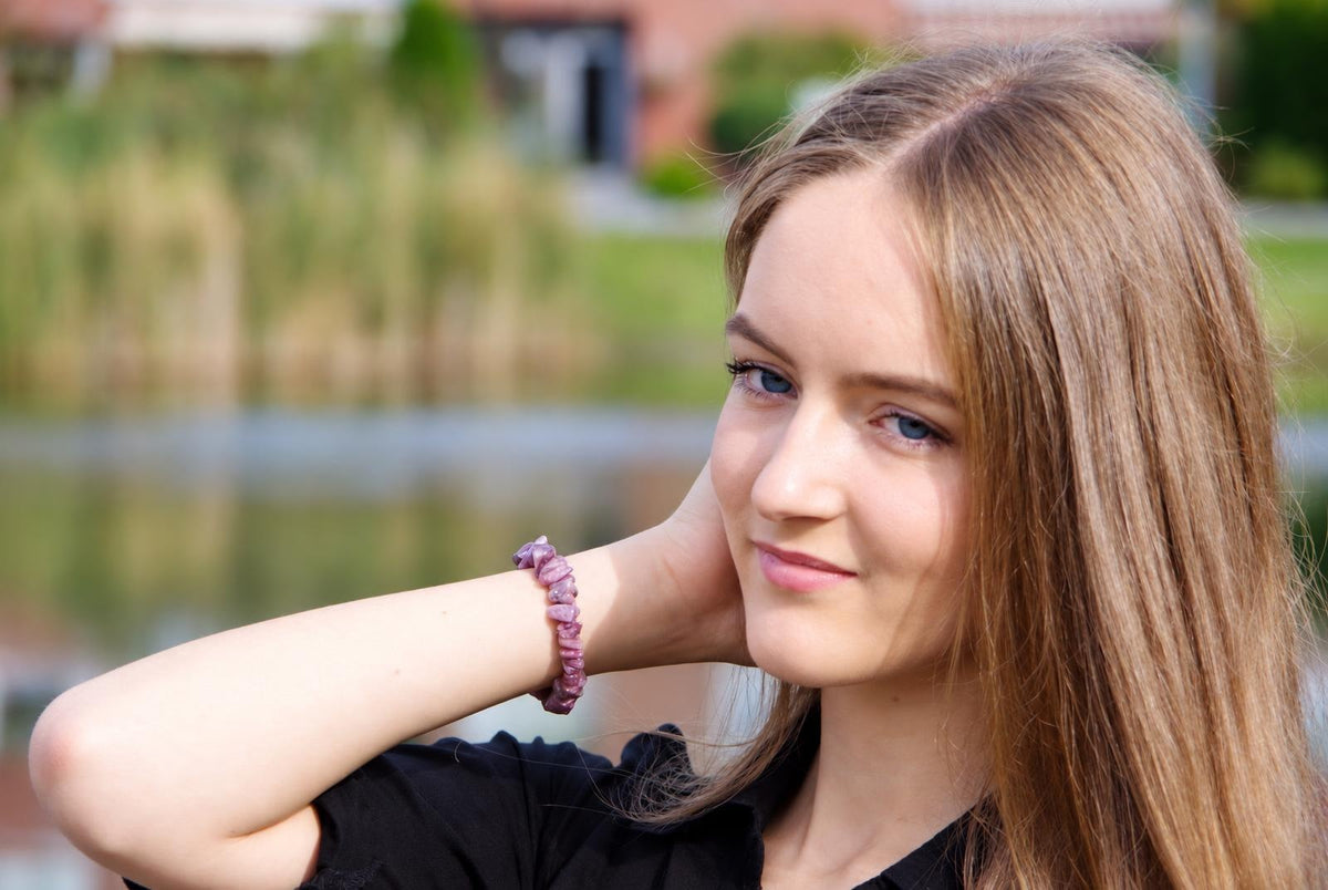Mergaitė su ilgomis šviesiai raudonomis plaukais, dėvi juodą marškinėlę ir violetinę akmenų apyrankę ant rankos.