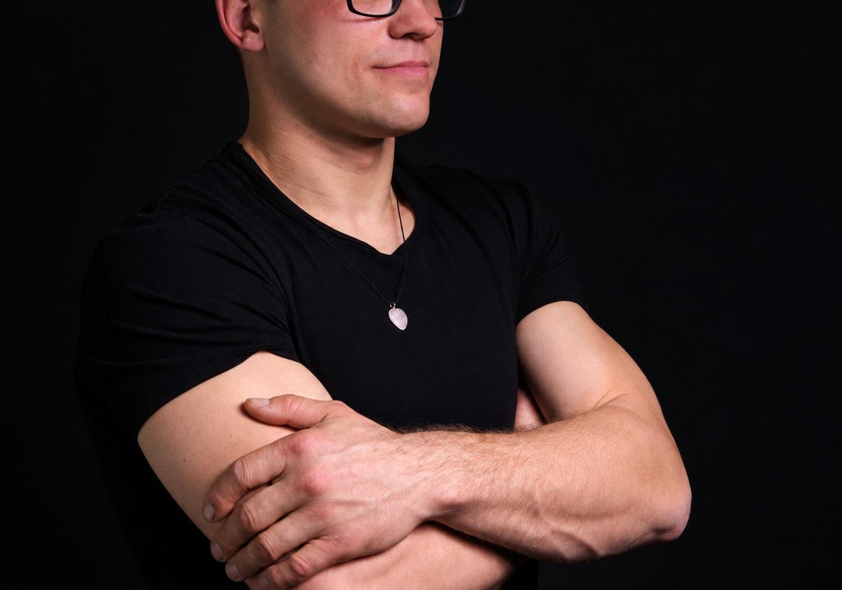 Vyras juodu marškinėliu dėvi kaklo apyrankę su rožinio kvarco širdies formos pakabuku ant tamsaus fono.