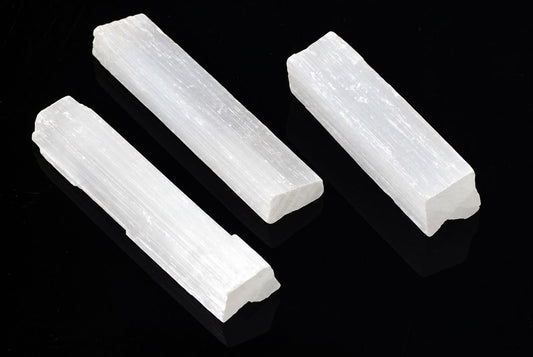 Trys baltos, plokščios selenito lazdelės ant tamsaus paviršiaus.