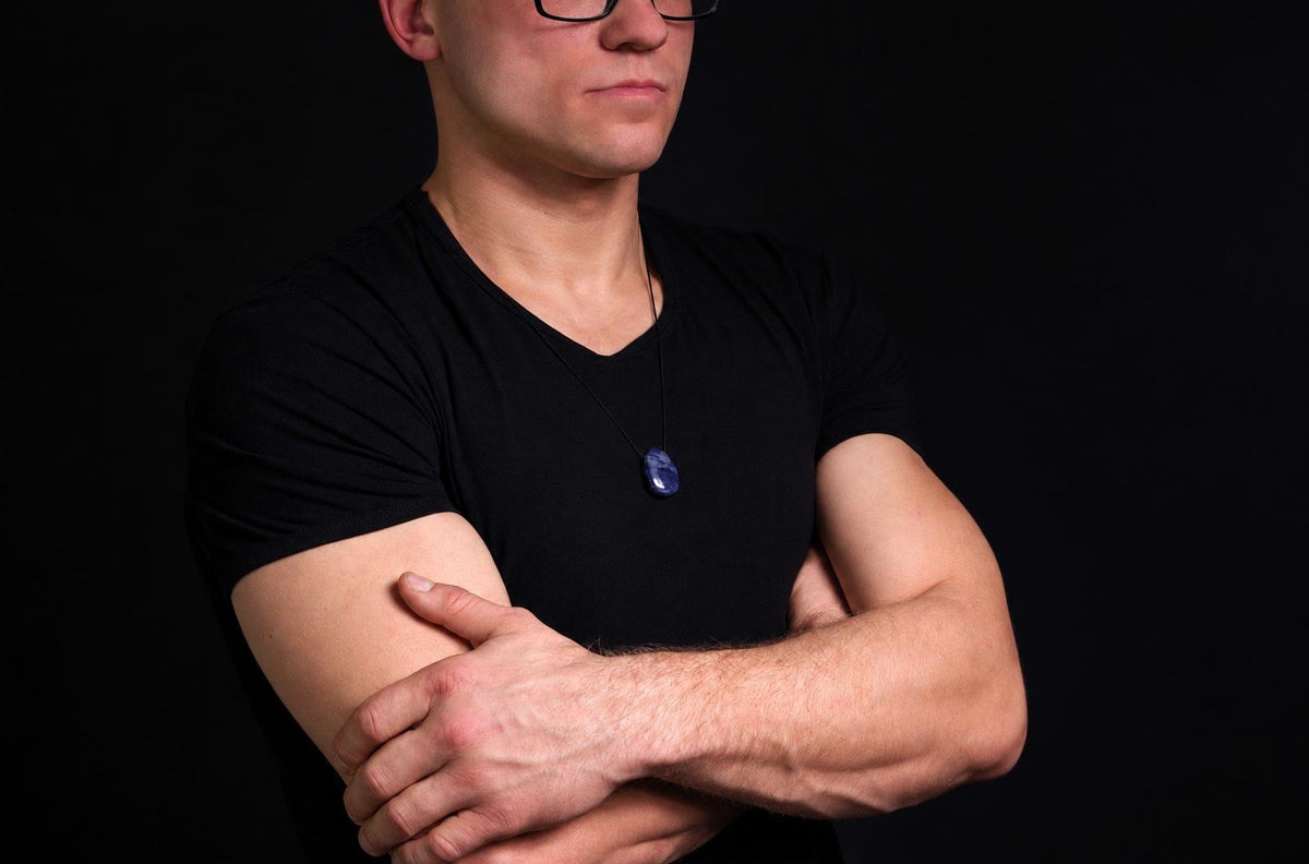 Vyras juodu marškinėliu dėvi pakabuką su tamsiai mėlynu, ovaliu akmenu ant kaklo prieš tamsų foną.