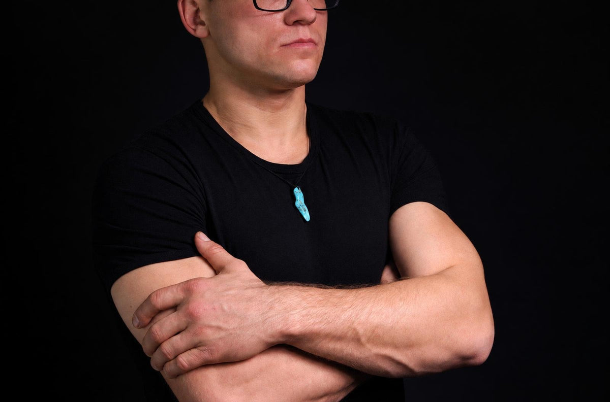 Vyras juodu marškinėliu dėvi tamsiai mėlyną, netaisyklingos formos pakabuką ant kaklo prieš tamsų foną.