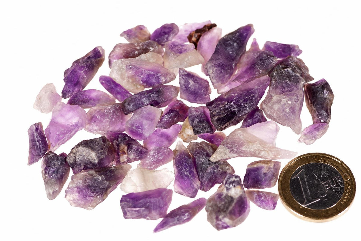 Baltame fone išdėstyti ametozo gabalėliai su violetnomis ir baltomis dalimis, šalia – vieno euras monetos.