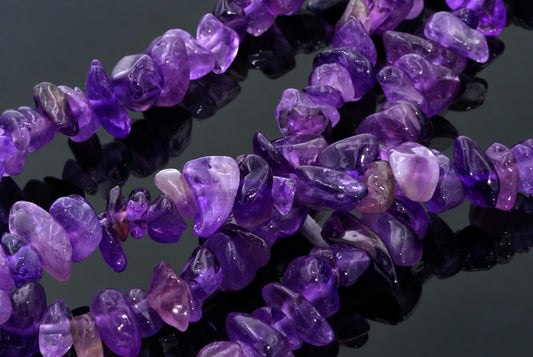 Užrašyti violetni ametoistų karoliai su netaisyklinga forma, išdėstyti ant tamsaus paviršiaus.