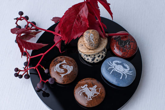 Juodas, blizgus diskas su penkiais apvalakais akmenimis, ant kurių balti zodiako ženklai. Raudonos lapų šakos ir uogos.