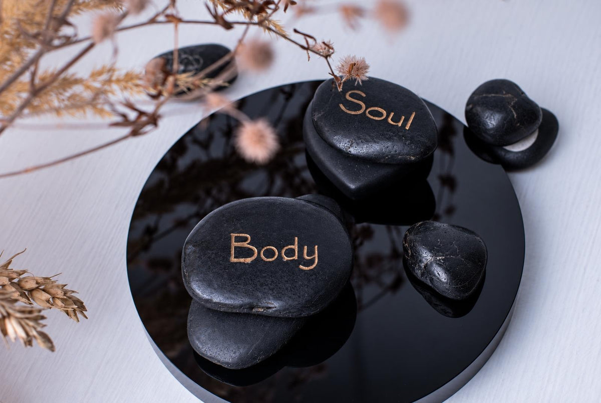 Juodi akmenys su išgraviruotais užrašais „Body“ ir „Soul“ ant juodo atspindinčio paviršiaus, šalia sausų augalų.
