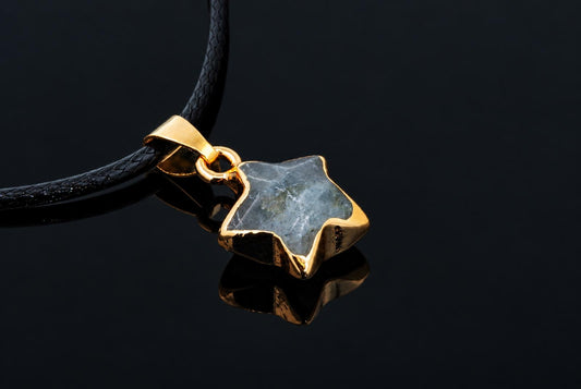 Auksinis pakabukas su žvaigždės formos larvikito akmeniu ant juodos odos grandinės, atspindintis tamsiame paviršiuje.