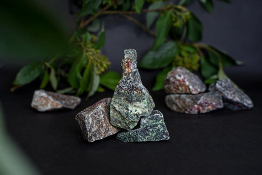 Keli žalios, rudos ir rausvos spalvos mineralų uolienos ant tamsaus paviršiaus su žaliais lapais fone.