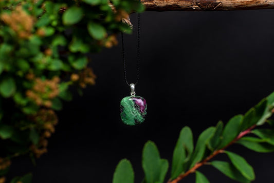 Pakabukas su žalios ir violetinės spalvos akmeniu, sidabrinėmis detalėmis, ant juodos grandininės, prieš tamsų foną.