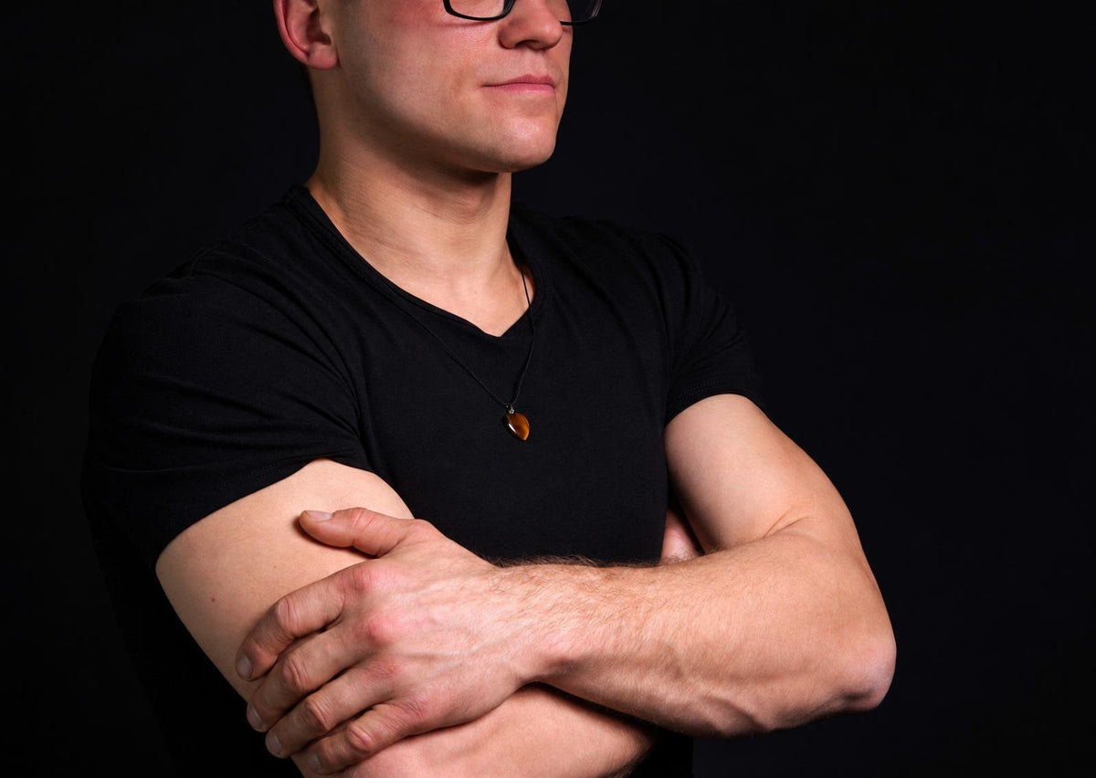 Vyras juodu marškinėliu dėvi kaklo papuošalą su rudojo atspalvio širdies formos pakabuku ant tamsaus fono.