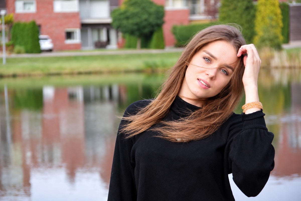 Mergina juodame viršutiniame drabužyje prie vandens, su šviesiai ruda plaukų ir oranžine rankinė.