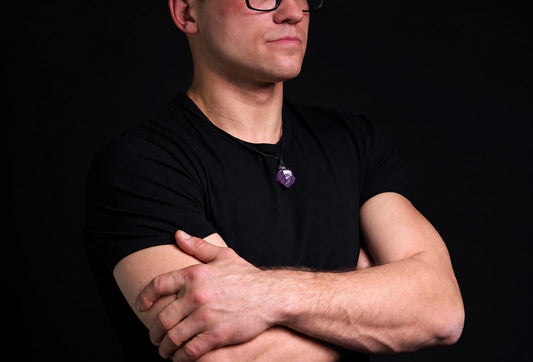 Vyras juodu marškinėliu dėvi kaklo pakabuką su violetiniu, neapdirbtu akmeniu ant tamsaus fono.