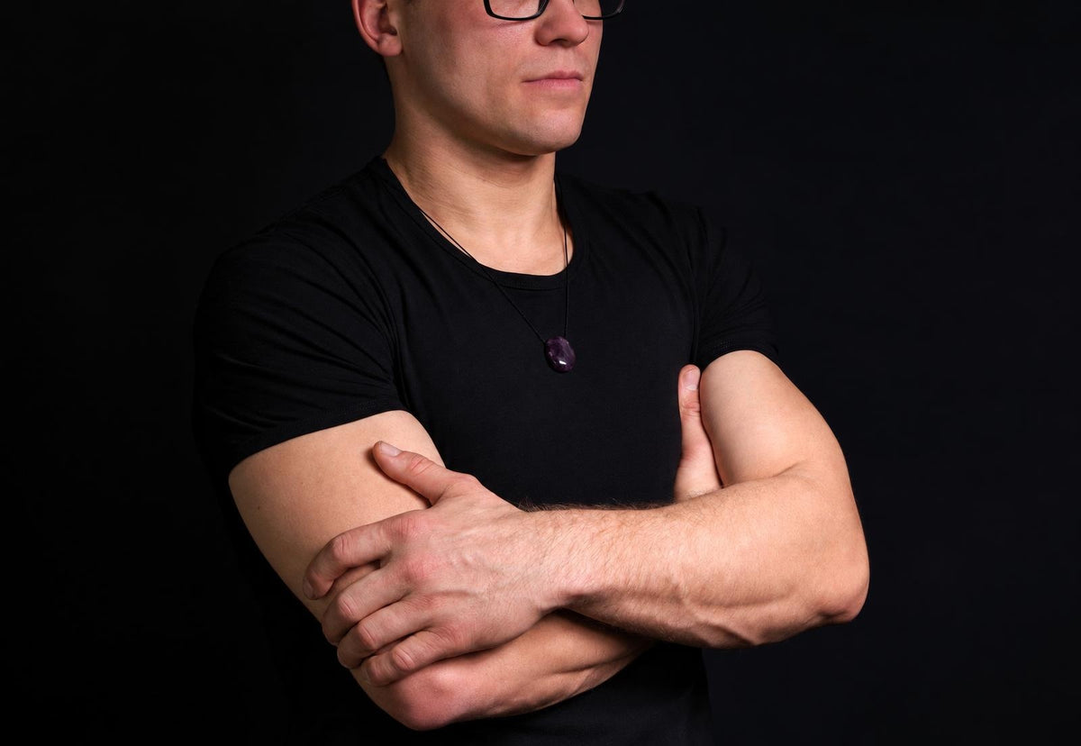 Vyras juodu marškinėliu su kaklo apyranka, turinčia tamsiai violetinį, lašo formos akmenį ant juodos grandinės.