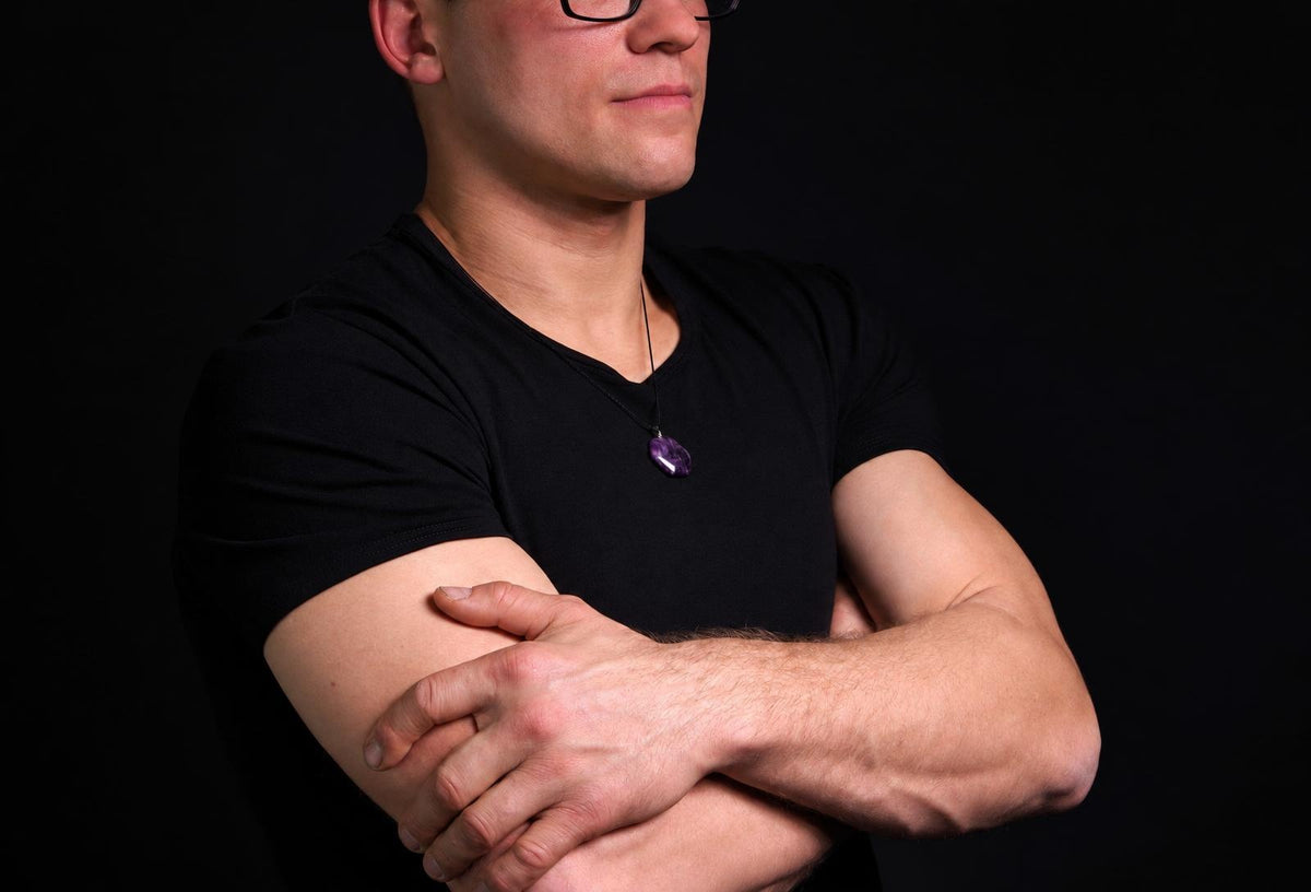 Vyras juodu marškinėliu dėvi kaklo apyrankę su violetiniu, šešiakampiu akmeniu ant tamsaus fono.