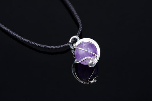 Pakabukas su violetva sfera, apvyniota sidabro spalvos metaline siūleliu, ant tamsaus paviršiaus.