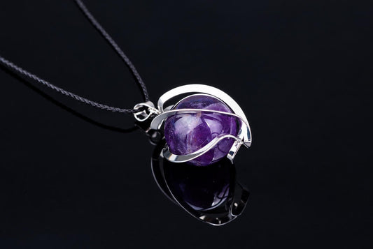 Pakabukas su violetva sfera, apvyniota sidabro spalvos metaline juosta, ant tamsaus paviršiaus.