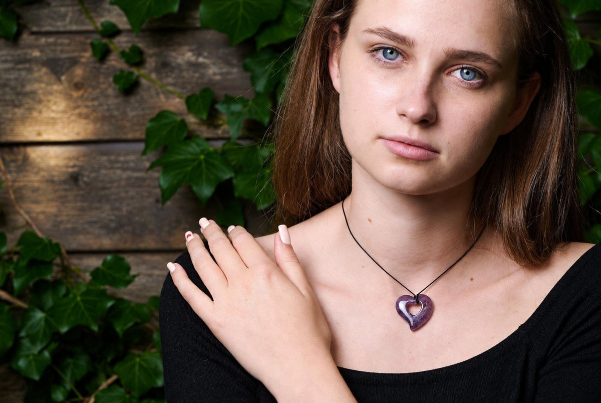 Moteris dėvi juodą kaklaraištį su violetiniu širdies formos pakabuku ant tamsaus fono su žaliais lapais.