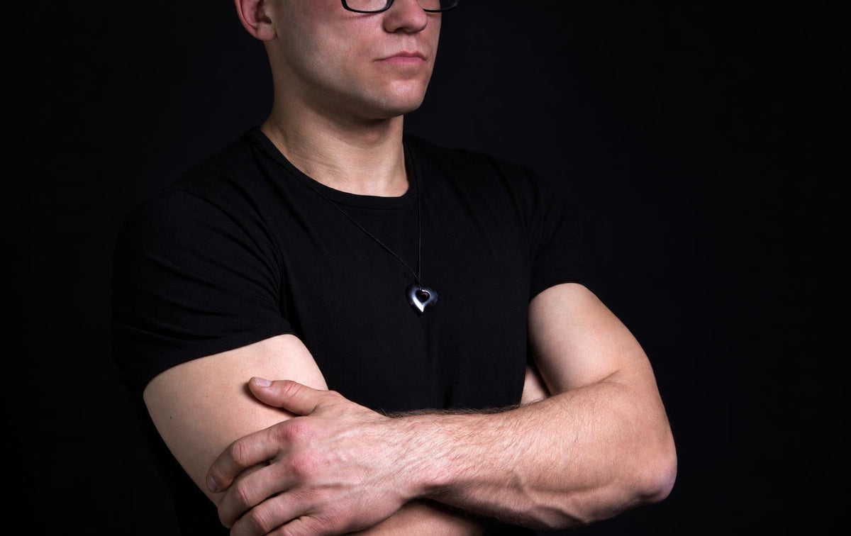 Vyras juodu marškinėliu dėvi širdies formos pakabuką ant kaklo, stovėdamas su kryžtomis rankomis prieš tamsų foną.