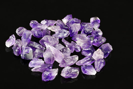Pile of rough amethyst crystals in shades of purple and clear on a dark surface.