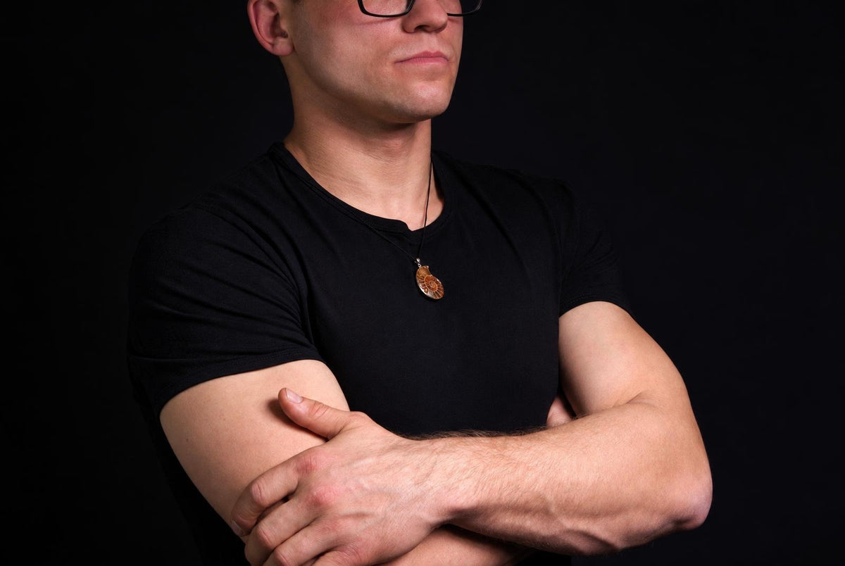 Vyras juodu marškinėliu dėvi pakabuką su amonitu ant kaklo, stovėdamas prieš tamsų foną.