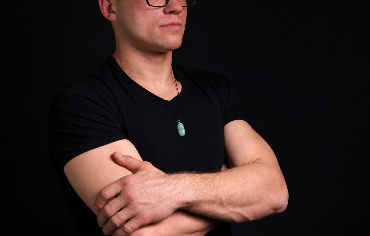 Vyras juodu marškinėliu dėvi kaklo pakabuką su šviesiai žaliu, ovaliu akmenuku ant tamsaus fono.