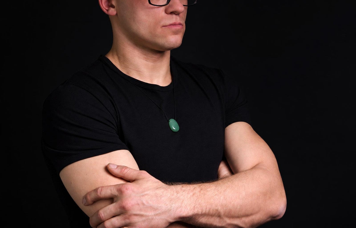 Vyras juodu marškinėliu dėvi pakabuką su žaliu, lašo formos akmeniu ant tamsaus fono.