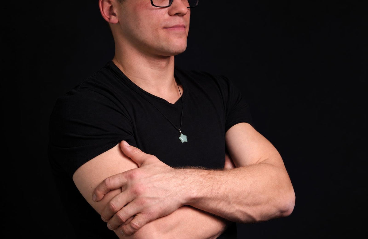 Vyras juodu marškinėliu dėvi žalią žvaigždės formos pakabuką ant kaklo, stovėdamas su kryžtomis rankomis prieš tamsų foną.