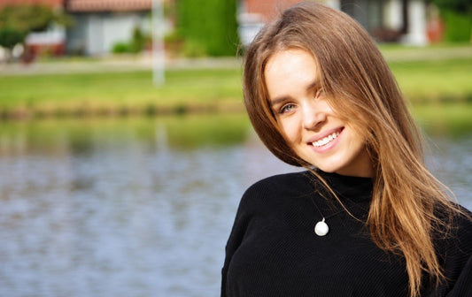 Mergina juodame viršutiniame drabužyje dėvi baltą apvalų pakabuką ant kaklo, stovėdama prie vandens.