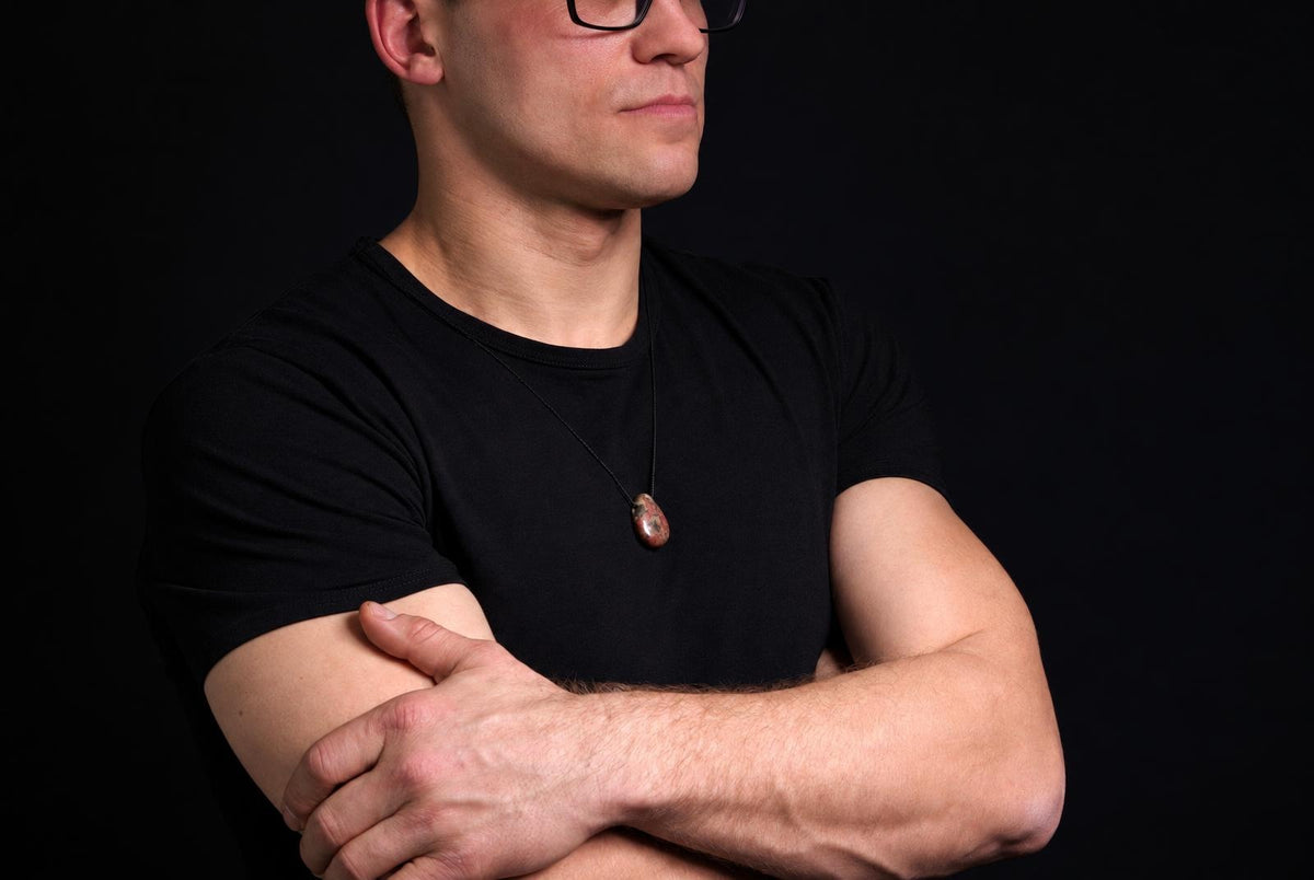 Vyras juodu marškinėliu dėvi kaklo pakabą su raudonai ruda, ovališka brekčijos jaspio akmeniu ant tamsaus fono.