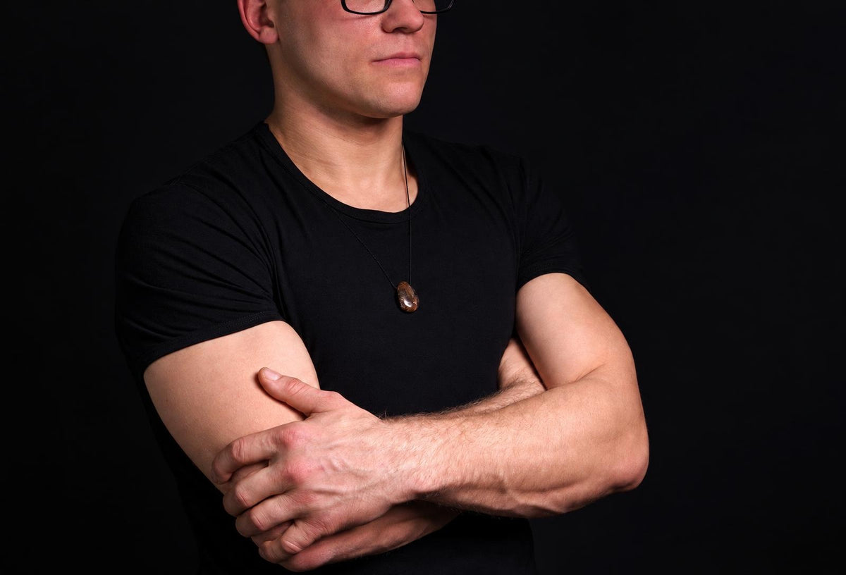 Vyras juodu marškinėliu dėvi pakabuką su rudojo atspalvio, ovalo formos akmeniu ant tamsaus fono.