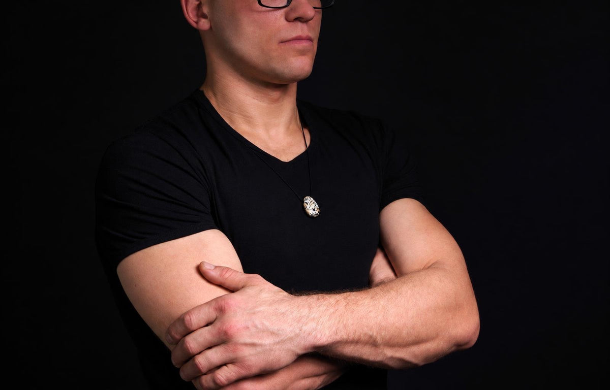 Vyras juodu marškinėliu dėvi kaklo pakabą su ovaliu, baltu ir tamsiai dėmėtu akmeniu ant tamsaus fono.