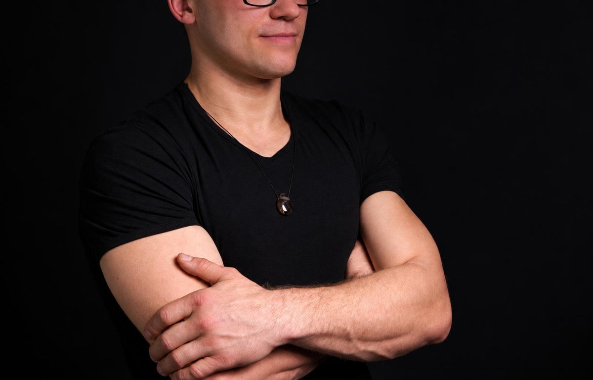 Vyras juodu marškinėliu dėvi kaklo apyrankę su tamsiai ruda, lašo formos pakabuka ant juodos fono.