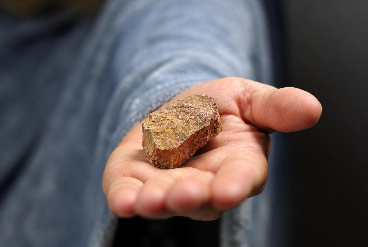 Žmogaus rankoje, ant tamsaus fono, matomas neapdirbtas rudos ir geltonos spalvos akmuo su šiurkščia tekstūra.