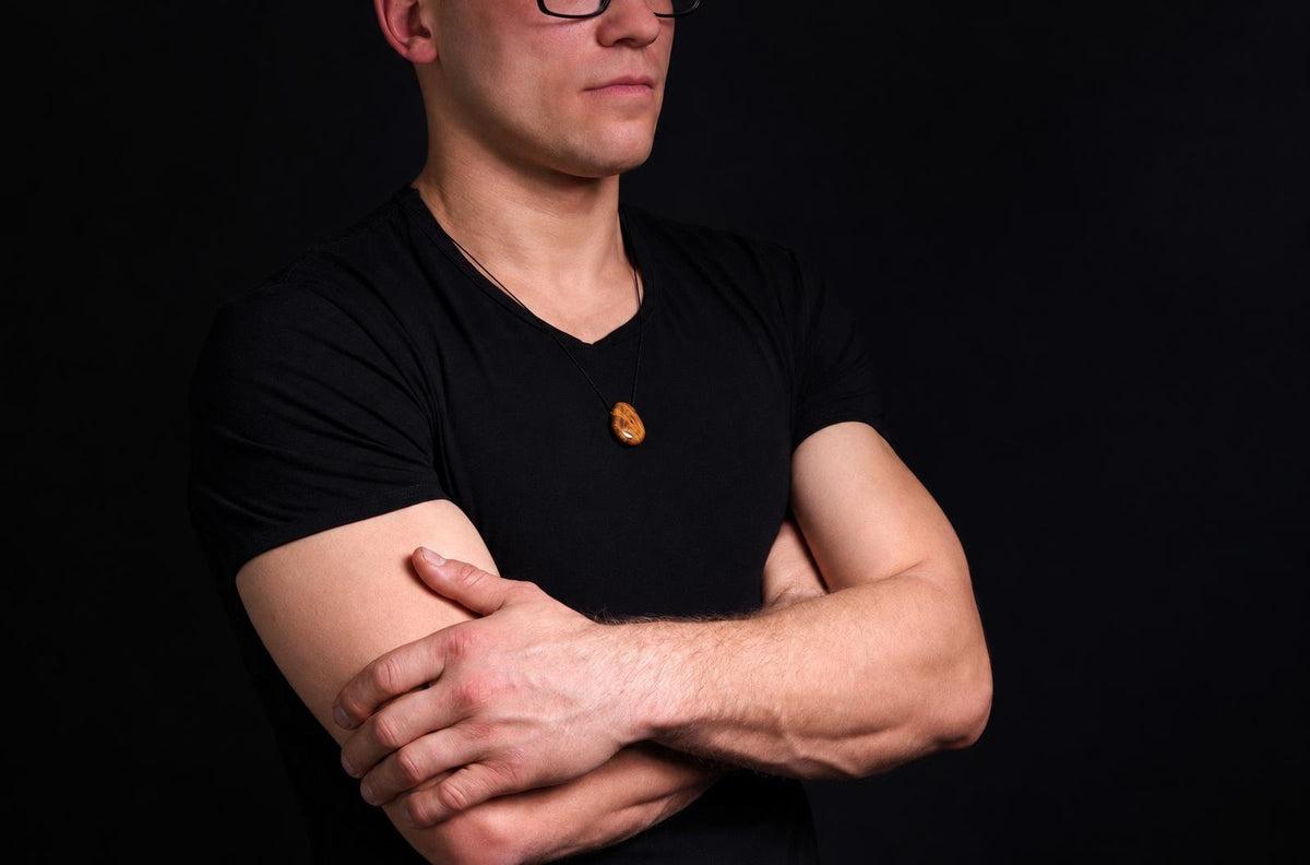 Vyras juodu marškinėliu dėvi kaklo apyrankę su ovaliu, rudojo atspalvio akmeniu ant tamsaus fono.