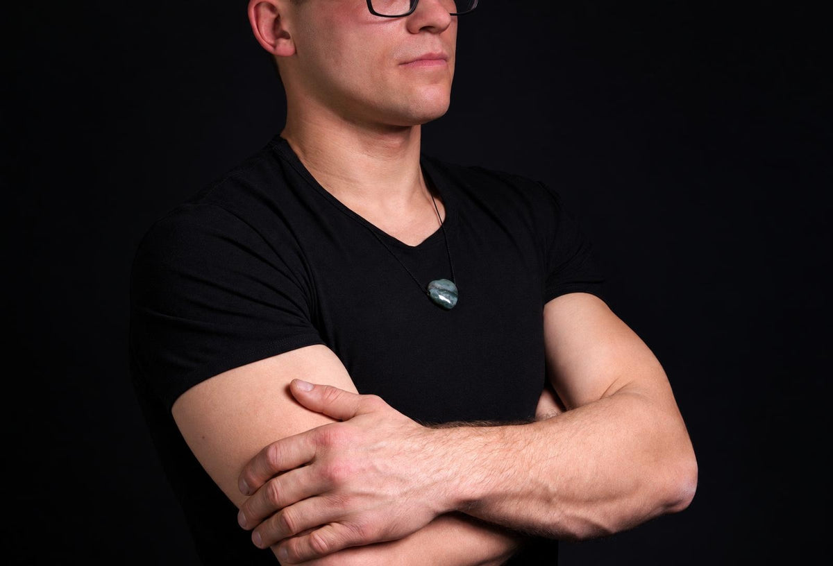 Vyras juodu marškinėliu dėvi pakabuką su tamsiai žaliu, apvaliu akmeniu ant kaklo.