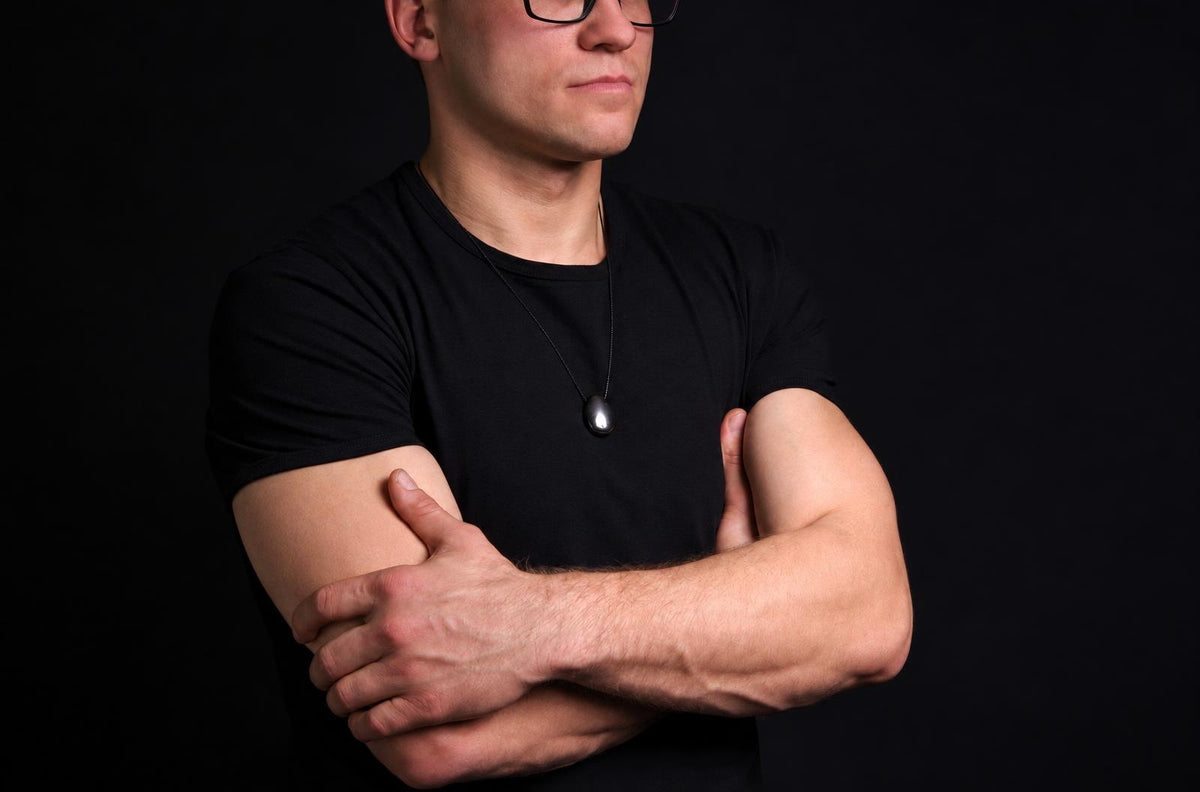 Vyras juodu marškinėliu dėvi pakabuką su tamsiai pilku, lašo formos akmeniu ant kaklo. Fonas – juoda.