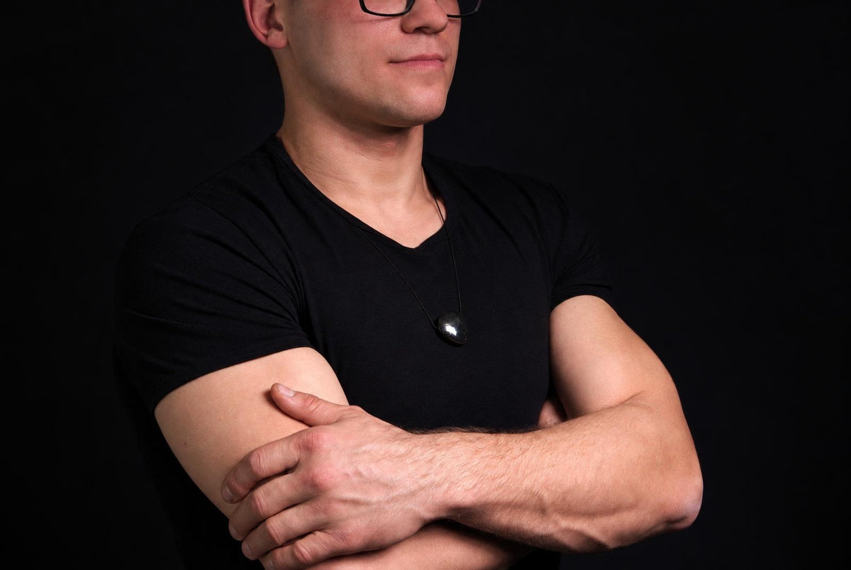 Vyras juodu marškinėliu dėvi pakabuką su tamsiai pilku, apvaliu akmeniu ant kaklo. Fonas tamsus.