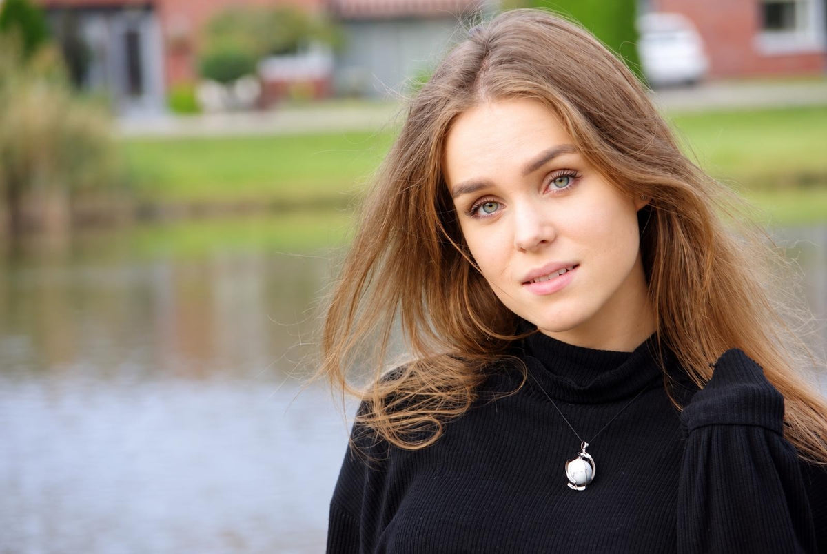 Mergina juodos megdėgės su sidabro spalvos sferiniu pakabuku ant kaklo, stovinti prie vandens.