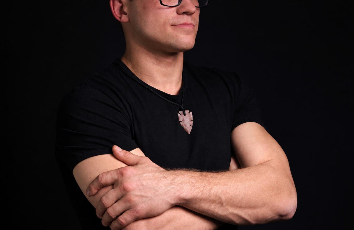 Vyras juodu marškinėliu dėvi šviesiai rausvos spalvos, strėlės formos pakabuką ant kaklo prieš tamsų foną.