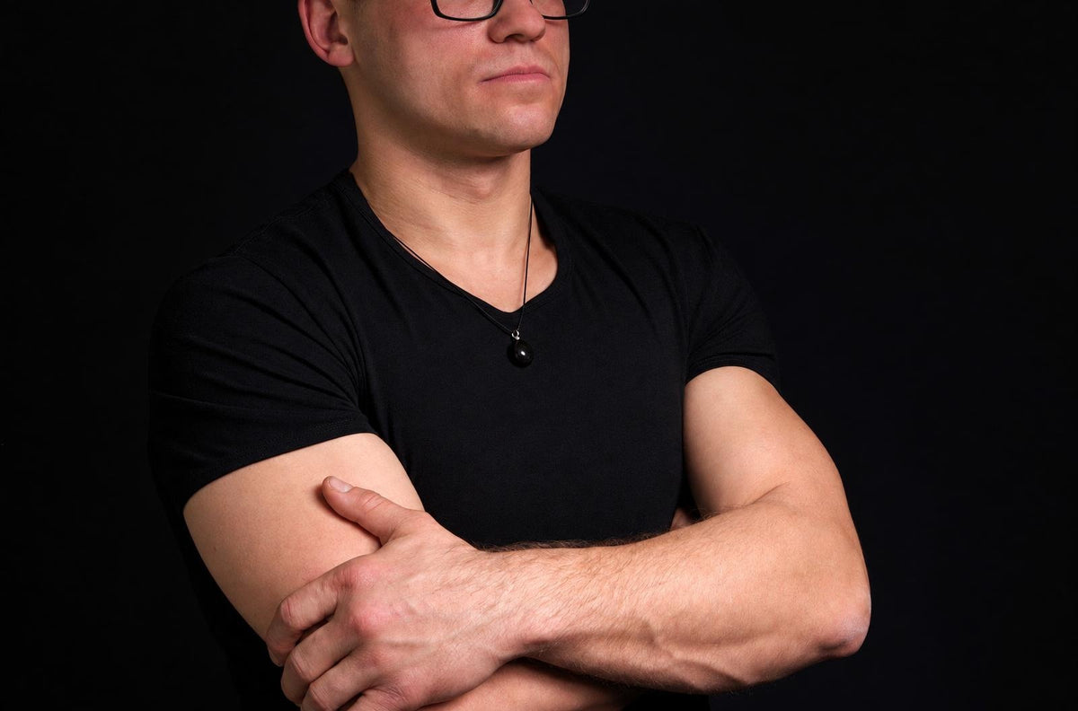 Vyras juodu marškinėliu su akmeniniu pakabuku kakle, stovintis prieš tamsų foną.