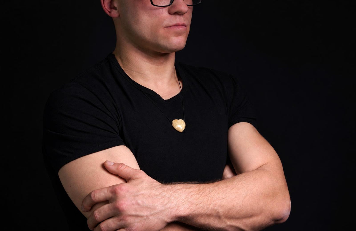 Vyras juodu marškinėliu dėvi kaklo papuošimą su šviesiai geltona, širdies formos pakabuka ant tamsaus fono.