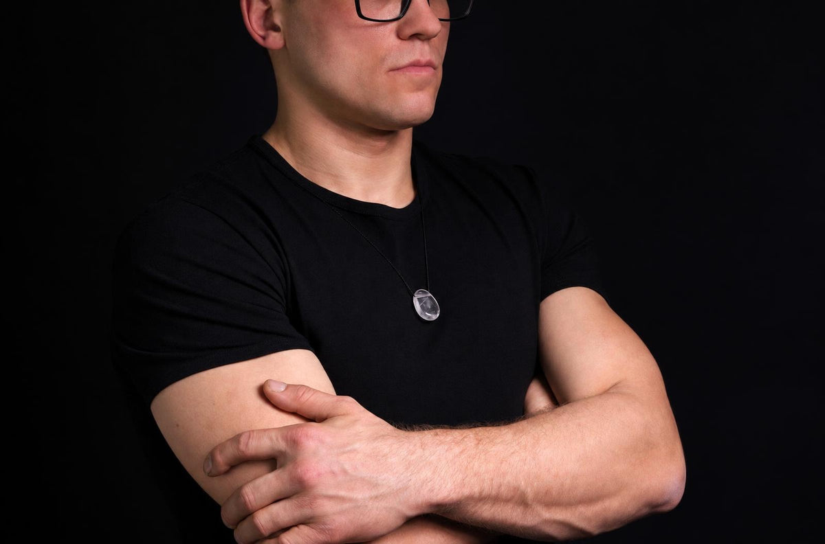 Vyras juodu marškinėliu dėvi pakabuką su skaidriu, lašo formos akmeniu ant tamsaus fono.